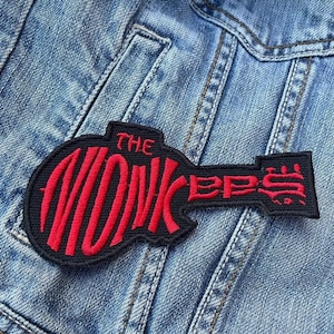 May include: A black and red embroidered patch in the shape of a guitar, with the words "THE MONKEES" in red. The patch is sewn onto a light blue denim jacket.