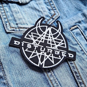 May include: Black and white embroidered patch with the word "DISTURBED" and a stylised logo featuring a circle with a star inside and two horns on top.
