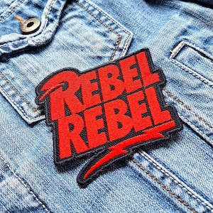Op de afbeelding: Rode en zwarte geborduurde patch met de tekst "Rebel Rebel" op een denim achtergrond.