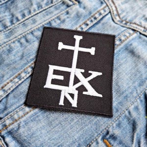 Könnte beinhalten: Schwarzer Patch mit weiß gesticktem Kreuz und den Buchstaben "EXN" auf einem Jeans-Hintergrund.