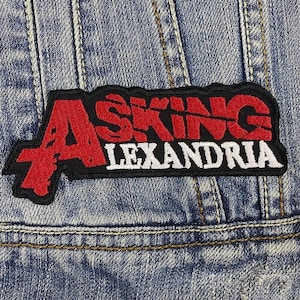 Könnte beinhalten: Ein rechteckiger Aufnäher mit den Worten "ASKING ALEXANDRIA" in roter und weißer Schrift auf schwarzem Hintergrund. Der Aufnäher ist auf eine blaue Jeansjacke genäht.