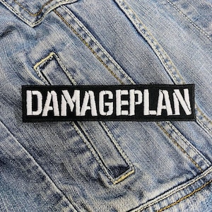 Puede incluir: Un parche negro y blanco con el texto "DAMAGEPLAN" en letras blancas sobre fondo negro. El parche está cosido en una chaqueta vaquera azul.