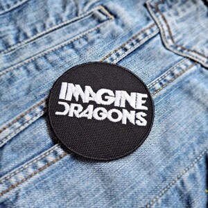 Op de afbeelding: Zwart-wit geborduurde patch met de tekst "Imagine Dragons" op een denim achtergrond.