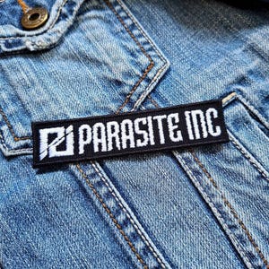 以下が含まれることがあります： デニムジャケットに縫い付けられた「PARASITE INC」の文字が入った、白と黒の長方形の刺繍パッチ。 パッチは白い文字と様式化されたロゴが特徴です。