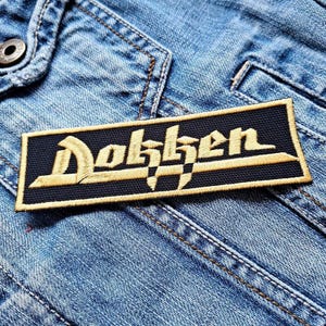 Op de afbeelding: Een rechthoekige patch met het woord "Dokken" geborduurd in goudkleurig garen op een marineblauwe achtergrond. De patch is genaaid op een lichtblauwe spijkerjas, met zichtbare stiksels en een metalen knoop.
