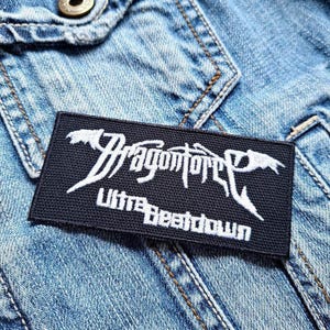 Pode incluir: Patch bordado preto e branco com o nome da banda "DragonForce" e as palavras "Ultra Beatdown" em uma jaqueta jeans.