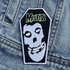 May include: A black and white Misfits patch with a skull design in a coffin shape. The word "Misfits" is in green lettering. The patch is sewn onto a denim jacket.