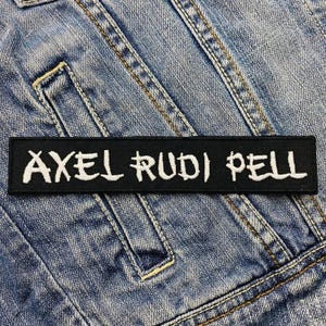 May include: A black rectangular patch with the white text "AXEL RUDI PELL". The patch is placed on a blue denim jacket. The text is in a stylized, handwritten font.