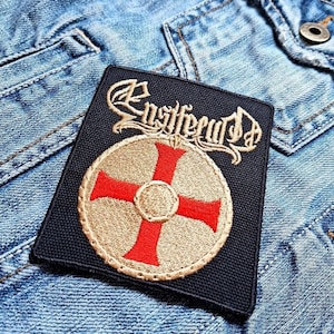 May include: Black embroidered patch with the band name "Enslaved" in white cursive lettering and a gold shield with a red cross in the center.