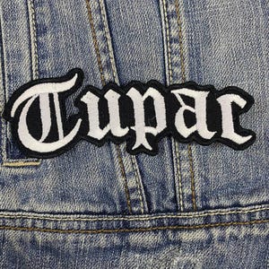 Op de afbeelding: Witte geborduurde patch met het woord "Tupac" in zwarte gotische letters op een blauwe denim achtergrond.
