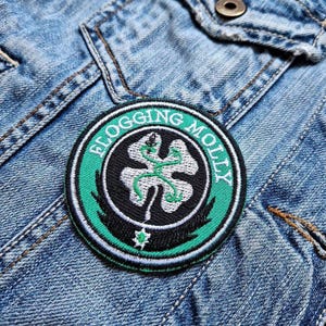 Peut inclure: Un patch brodé vert et blanc avec le texte "Flogging Molly" en cercle autour d'un trèfle à quatre feuilles.