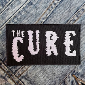 May include: Black embroidered patch with white lettering that reads "THE CURE".
