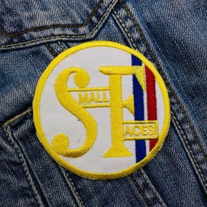 Puede incluir: Un parche circular blanco y amarillo con las letras "SF" en amarillo, "MALL" en texto negro sobre las letras y "ACES" en texto negro debajo de las letras. El parche tiene una franja azul, blanca y roja en el lado derecho.