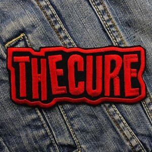 May include: Embroidered patch featuring the text "THE CURE" in bold red letters with a black border. The patch is rectangular and set against a blue denim background, showcasing the band's name.