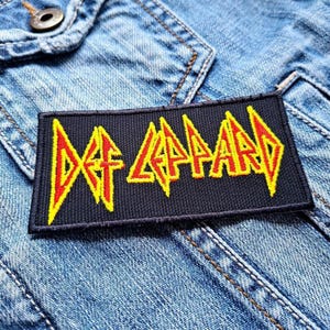 以下が含まれることがあります： 黒いアイロン接着パッチに、バンド名「Def Leppard」が黄色と赤の文字で書かれています。
