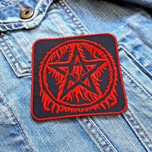 May include: Red pentagram with flames embroidered on a black patch. The patch is attached to a blue denim jacket.