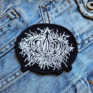 Op de afbeelding: Zwart-witte geborduurde patch met een gestileerd ontwerp op een zwarte achtergrond. Het ontwerp is rond met een complex patroon van lijnen en krommen.