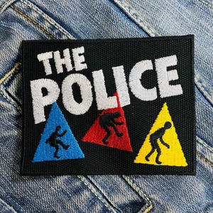 Può includere: Toppa ricamata nera con il testo "THE POLICE" in lettere bianche. Tre figure stilizzate si trovano negli angoli inferiori della toppa, ciascuna in un triangolo di colore diverso: blu, rosso e giallo.
