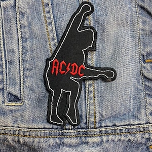 Könnte beinhalten: Schwarzer, gestickter Aufnäher mit weißem Rand, der eine stilisierte Figur und den roten Text "AC/DC" zeigt. Der Aufnäher ist auf eine Jeansjacke genäht.