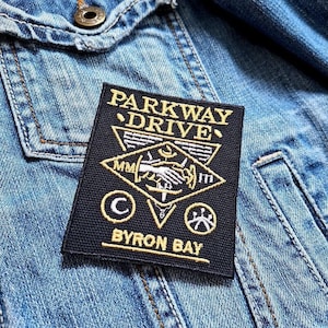 Op de afbeelding: Zwarte en gouden geborduurde patch met de tekst "Parkway Drive" en "Byron Bay". De patch heeft een gestileerde handdruk met een zwaard en andere symbolen.