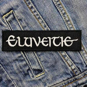 Op de afbeelding: Een zwart rechthoekig embleem met het woord "Eluveitie" in witte, gestileerde letters. Het embleem is geplaatst tegen een blauwe denim achtergrond, met zichtbare stiksels en naden. Het embleem is ontworpen om op kleding te worden genaaid.