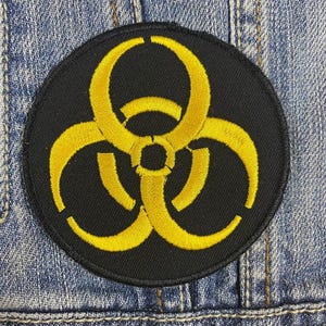 May include: A circular black patch featuring a yellow biohazard symbol. The symbol, a warning sign for biological hazards, is composed of three connected rings. The patch is designed to be sewn or ironed onto clothing or other fabric items.