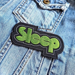 May include: A black and green embroidered patch with the word "Sleep" in a stylized font. The patch is attached to a blue denim jacket.