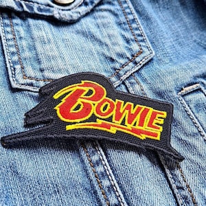 Peut inclure: Un écusson noir avec le nom "Bowie" en lettres rouges et jaunes, cousu sur une veste en jean. L'écusson a une forme unique avec un design stylisé.