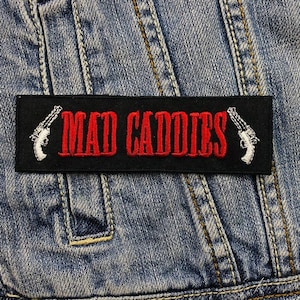 Może przedstawiać: Czarna naszywka haftowana z białymi pistoletami po obu stronach czerwonego napisu "MAD CADDIES".