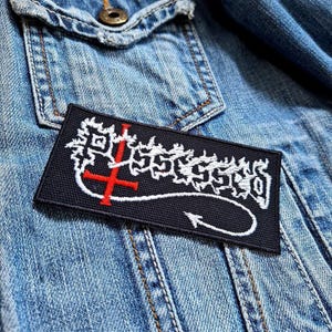 May include: Black and white embroidered patch with the word "Possessed" in a stylized font. A red inverted cross is above the word and a black tail with a white arrowhead extends from the right side of the word.
