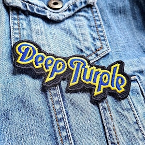 以下が含まれることがあります： 青いデニムジャケットに縫い付けられた、黒い刺繍パッチ。パッチには「Deep Purple」という文字が黄色と青の文字で刺繍されている。