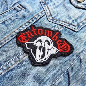May include: A black and white embroidered patch with the word "Entombed" in red lettering. The patch features a skull with a large, open mouth.