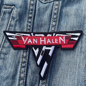 Può includere: Una toppa ricamata in bianco e nero con le parole "Van Halen" in rosso su uno stendardo bianco. La toppa ha la forma di una V stilizzata con le ali.