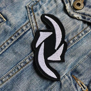 Puede incluir: Parche blanco bordado sobre fondo negro, con un diseño estilizado de dos flechas entrelazadas. El parche está sujeto a una chaqueta vaquera azul.