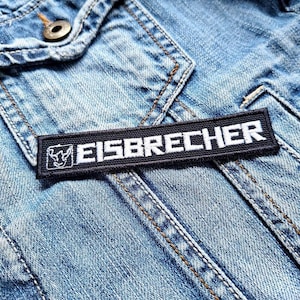 Könnte beinhalten: Ein schwarzer Patch mit weißen Nähten, auf dem "EISBRECHER" auf einer blauen Jeansjacke steht.