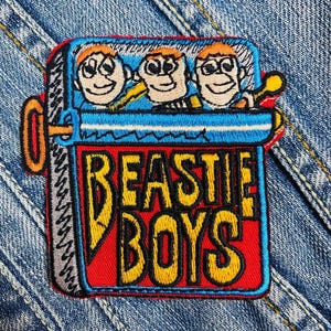 May include: A red, white, and blue embroidered patch with the words "Beastie Boys" in yellow letters. The patch features three cartoon faces inside a can.