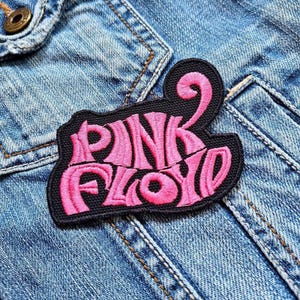 Könnte beinhalten: Ein schwarzer, gestickter Aufnäher mit den Worten "PINK FLOYD" in Pink, auf eine Jeansjacke genäht. Der Aufnäher hat eine einzigartige, stilisierte Schriftart und einen schwarzen Rand. Die Jeansjacke ist hellblau.