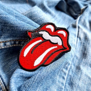 May include: Red and white embroidered patch of a tongue sticking out with a black outline. The patch is on a blue denim jacket.