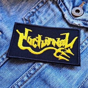 May include: A black patch with the word "Nocturnal" in yellow embroidered lettering. The lettering is stylized and has a gothic font. The patch is sewn onto a blue denim jacket.