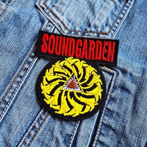 May include: A black and red embroidered patch with the text "SOUNDGARDEN" on a denim jacket. The patch is above a yellow and black embroidered patch with a spiral design.