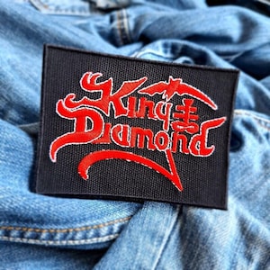May include: Black rectangular patch with the words "King Diamond" in red, stylized lettering. The patch is embroidered with a white outline and rests on a blue denim background. The patch is likely for a jacket or bag.