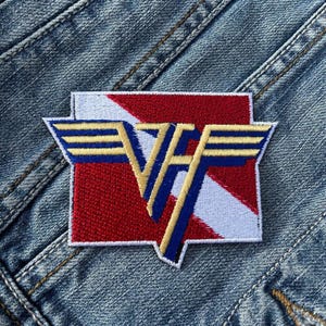 Op de afbeelding: Een geborduurde patch met een gestileerd "VF" logo met vleugels, in rood, wit en blauw. De patch is genaaid op een spijkerjack.
