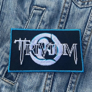 Puede incluir: Un parche bordado negro y azul con la palabra "Trivium" en letras blancas y un círculo blanco con un contorno azul.