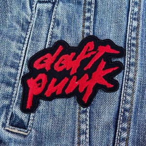Puede incluir: Parche negro con la inscripción roja "daft punk" cosido en una chaqueta vaquera azul. El parche es rectangular con esquinas redondeadas. La chaqueta vaquera tiene costuras visibles.