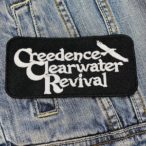 Könnte beinhalten: Schwarzer und weißer gestickter Aufnäher mit dem Text "Creedence Clearwater Revival" und einem stilisierten Vogelmotiv.