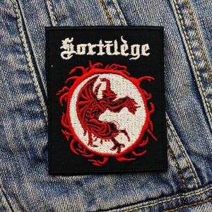 May include: Black embroidered patch with the band name "Sortilege" in white lettering and a red and white illustration of a winged creature in a circle.