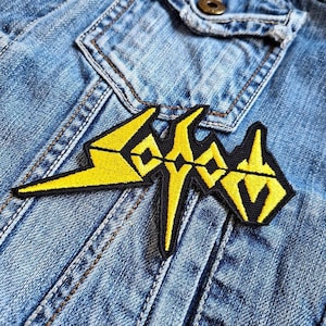 May include: A black and yellow embroidered patch featuring the word "SODOM" in a stylized font. The patch is sewn onto a light blue denim jacket. The patch has a lightning bolt design and is likely for a band or music-related merchandise.