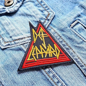 Peut inclure: Un écusson triangulaire noir avec les mots "Iron Maiden" en lettres jaunes et rouges. L'écusson est cousu sur une veste en jean bleue.