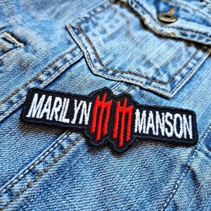 Puede incluir: Un parche bordado en blanco y negro con el texto "Marilyn Manson" y un símbolo estilizado rojo.