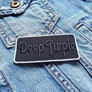 Op de afbeelding: Een zwart-wit geborduurde patch met de tekst "Deep Purple" op een blauwe denim jas.